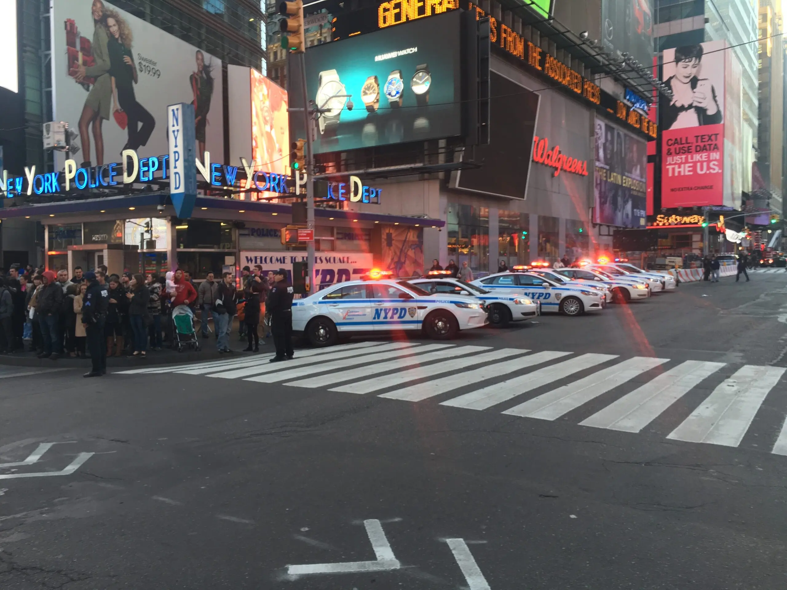 NYPD vehicles with flashing lights block a Times Square intersection, as a crowd gathers nearby, highlighting a significant police presence.