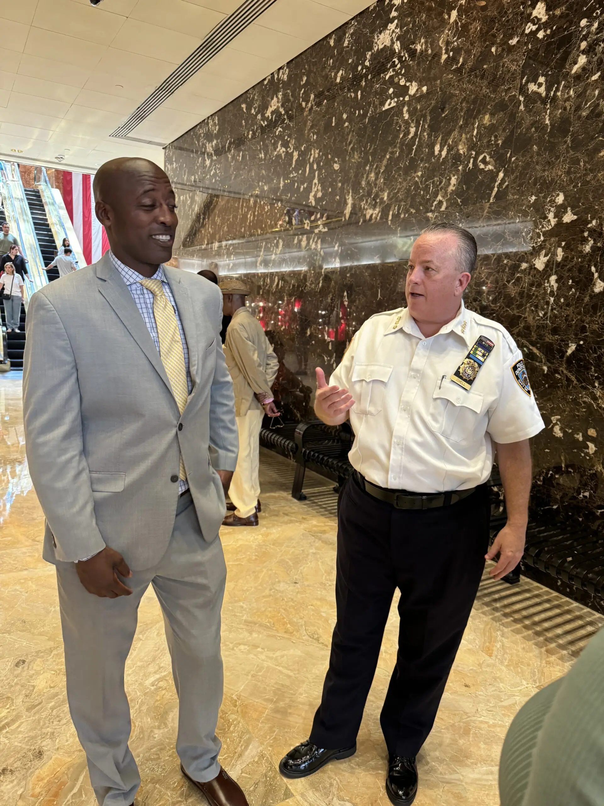Two men engage in conversation in a lobby, with one dressed in a light gray suit and the other in a police uniform.