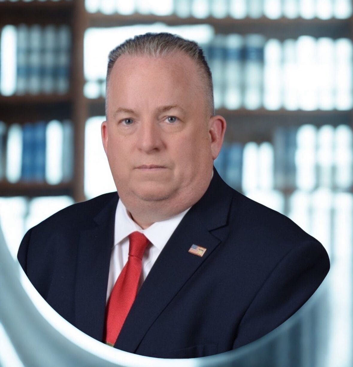 A man in a suit with a red tie poses confidently, showcasing an American flag lapel pin. A library backdrop hints at an academic or professional context.