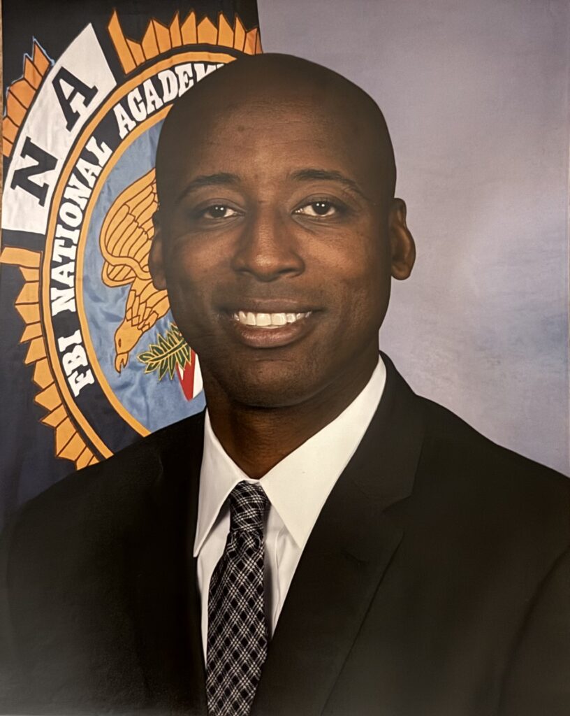Portrait of a smiling man in a suit, with the FBI National Academy emblem in the background, highlighting his affiliation with this prestigious institution.