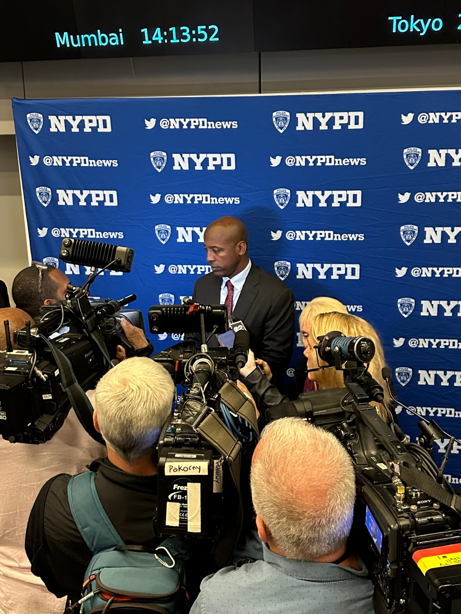 A police official addresses reporters surrounded by cameras at a press conference, with an NYPD backdrop and digital clocks displaying time in different cities.