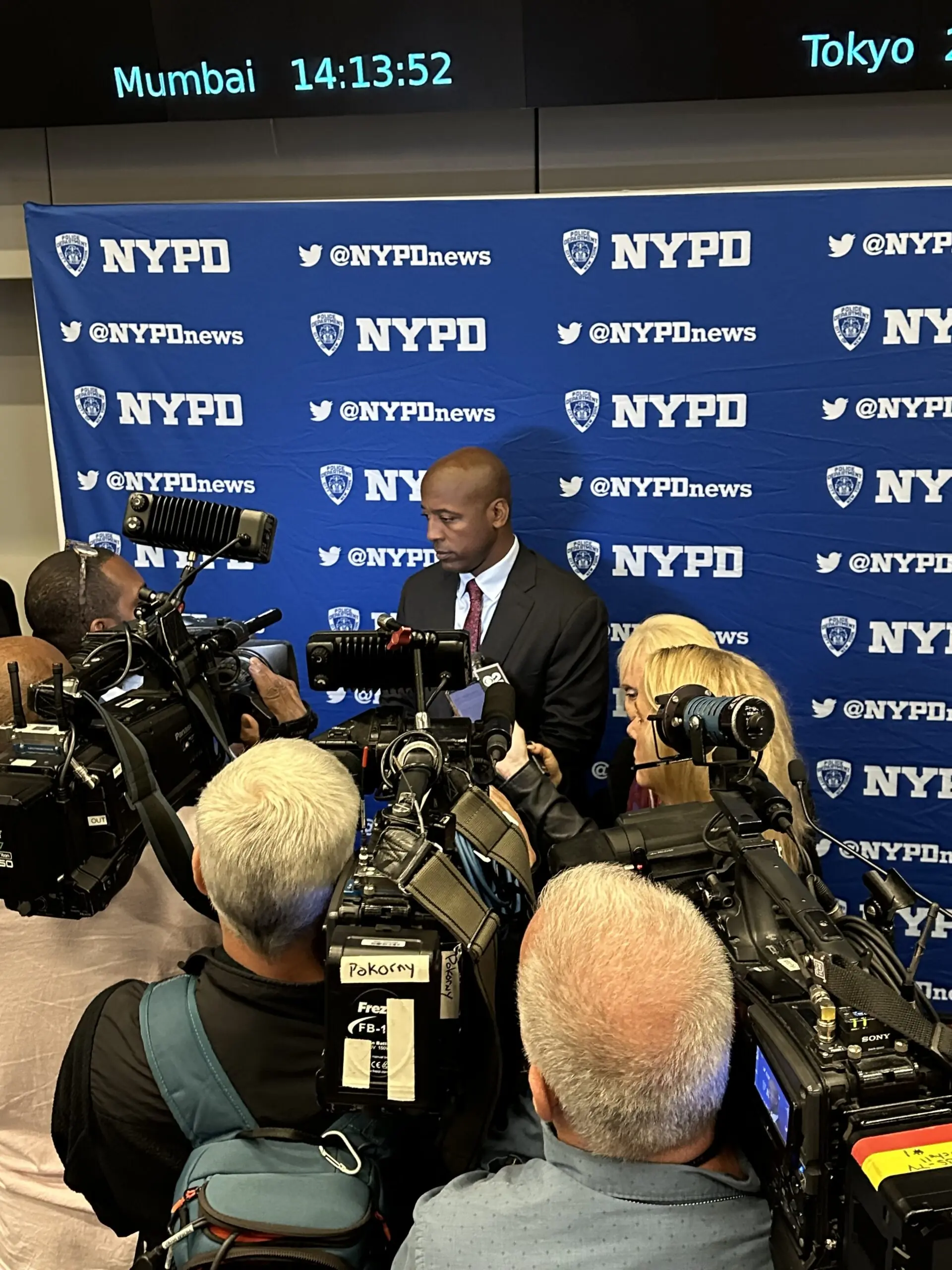 A police official addresses reporters surrounded by cameras at a press conference, with an NYPD backdrop and digital clocks displaying time in different cities.