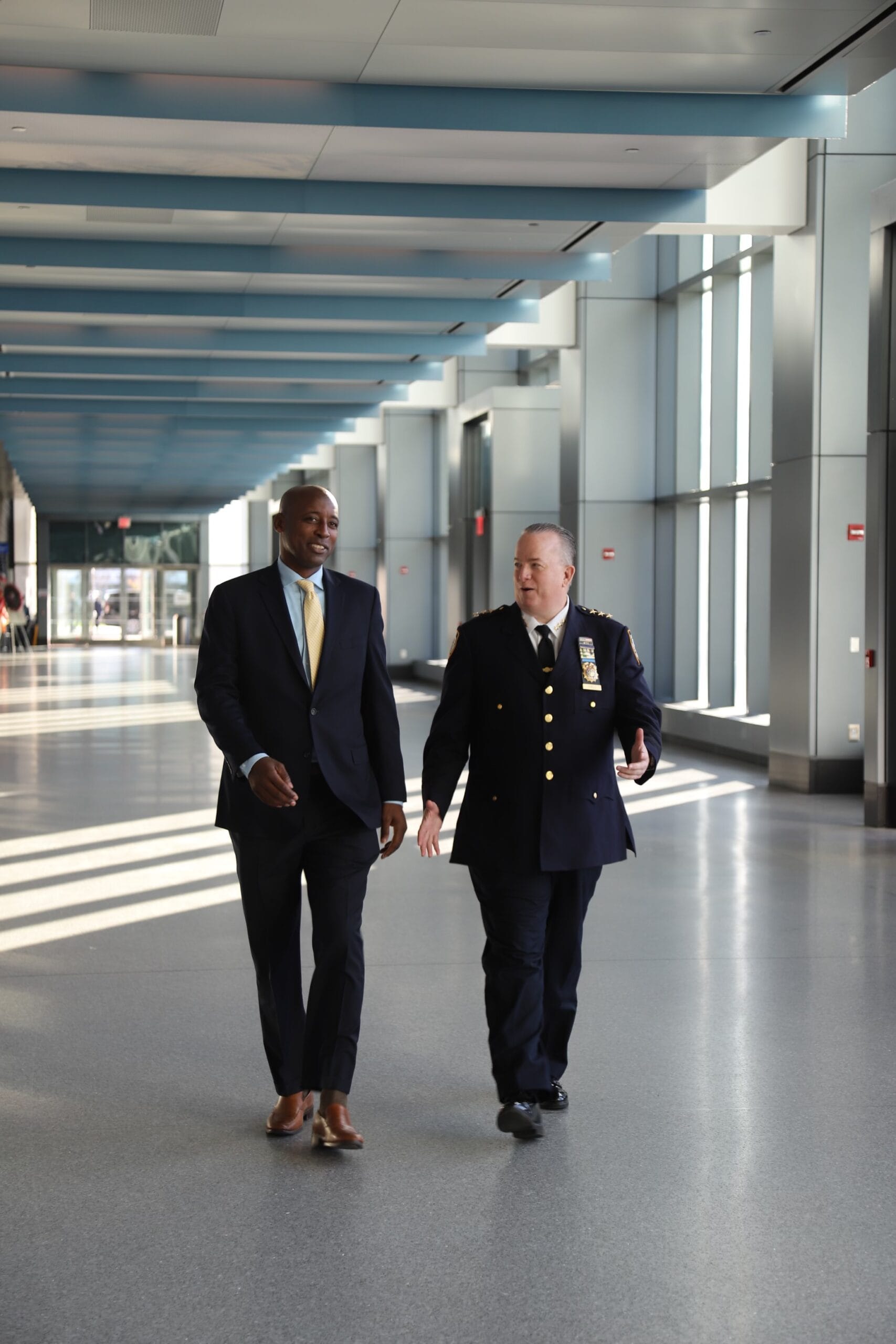 Two men, a business professional in a suit and a police officer in uniform, walk together through a spacious, sunlit corridor, discussing matters.