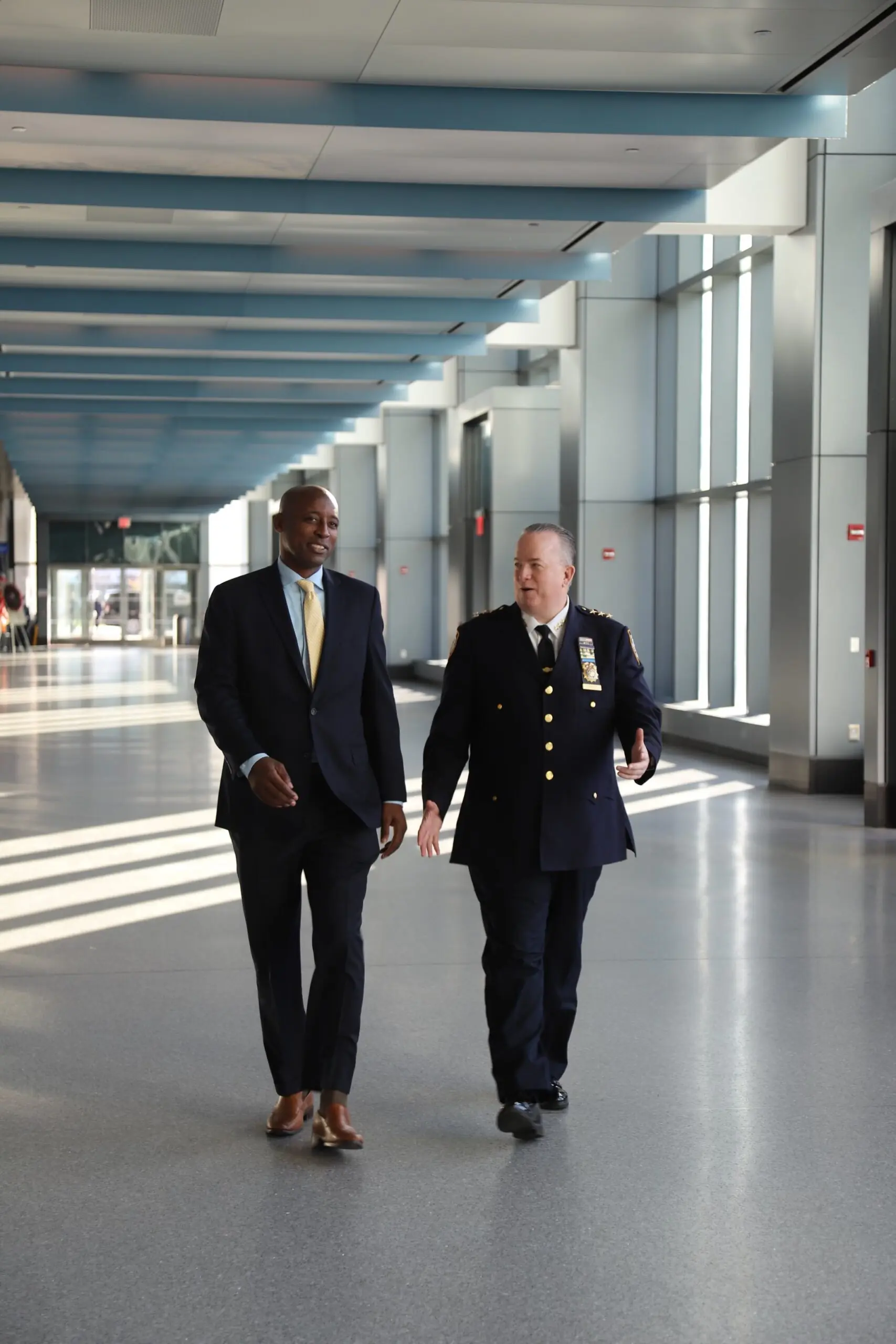Two men, a business professional in a suit and a police officer in uniform, walk together through a spacious, sunlit corridor, discussing matters.