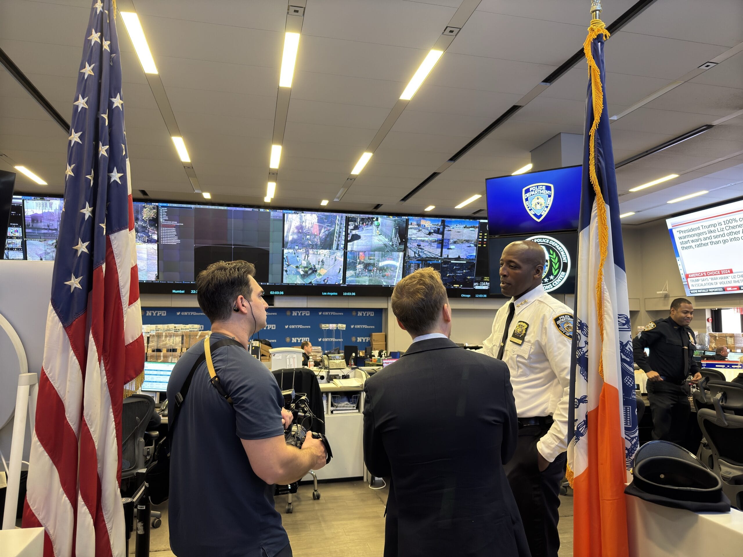 NYPD command center bustling with activity. An officer talks to two reporters, while security footage is displayed on large screens.