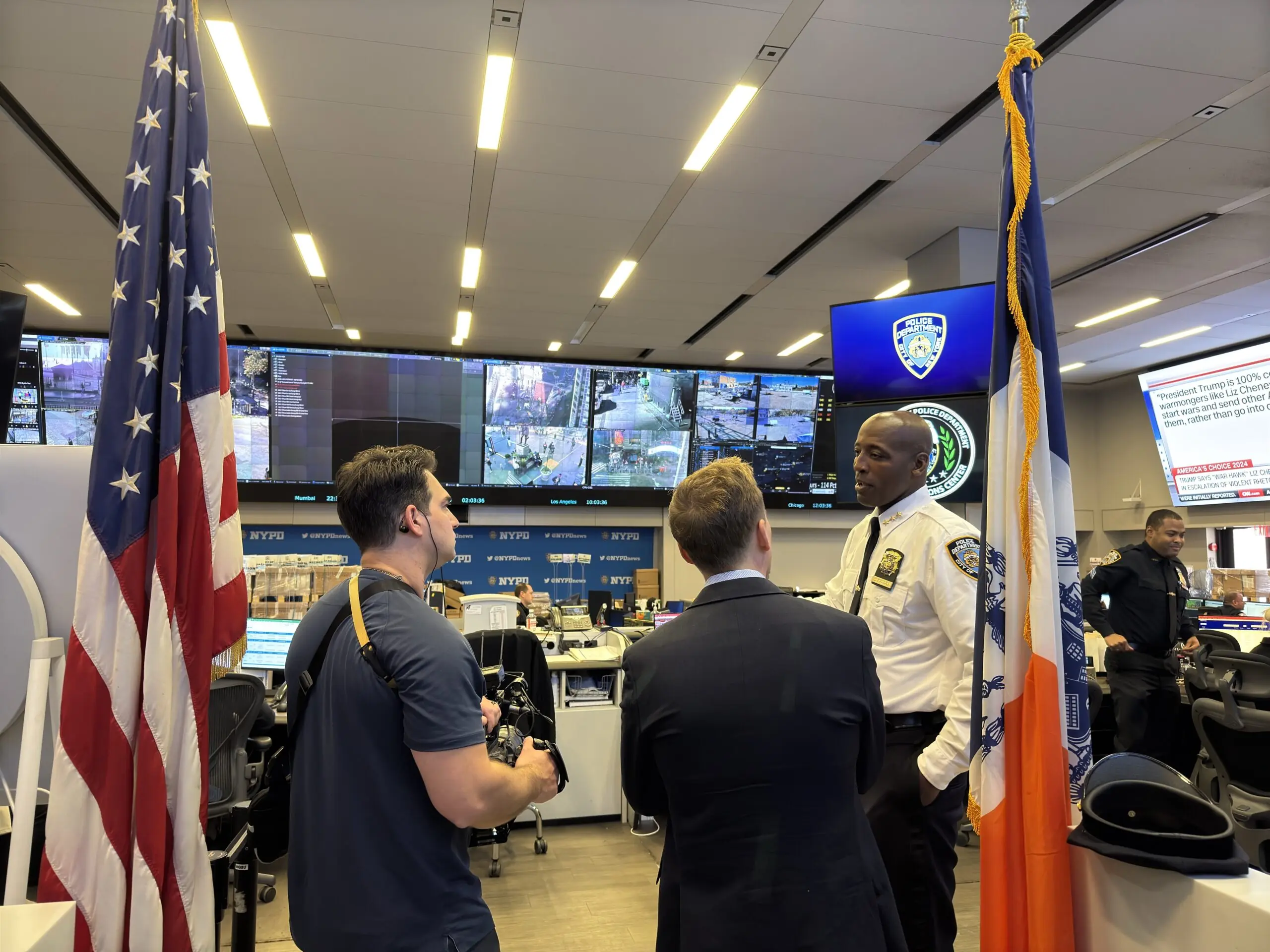 NYPD command center bustling with activity. An officer talks to two reporters, while security footage is displayed on large screens.